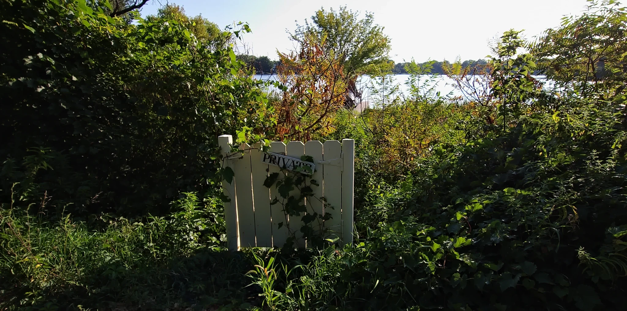 A wooden gate with a sign: "PRIVATE." The gate is in vegetation, unattached to any fence, and beyond it lies a lake.